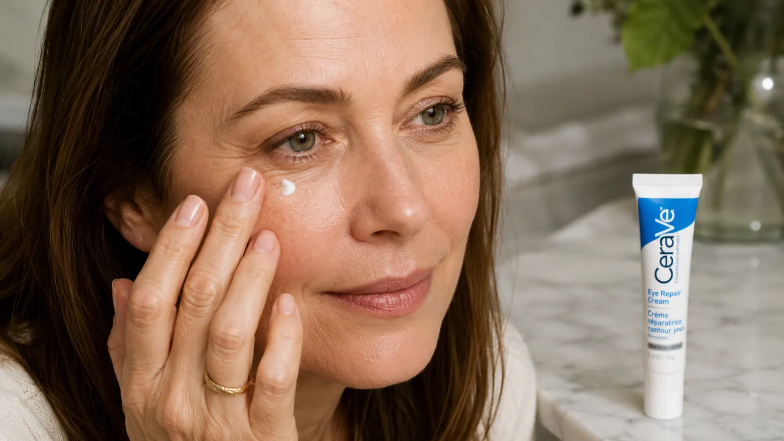 Close-up of a ring finger dabbing a small amount of eye cream near the under-eye area, tube resting on a bathroom counter