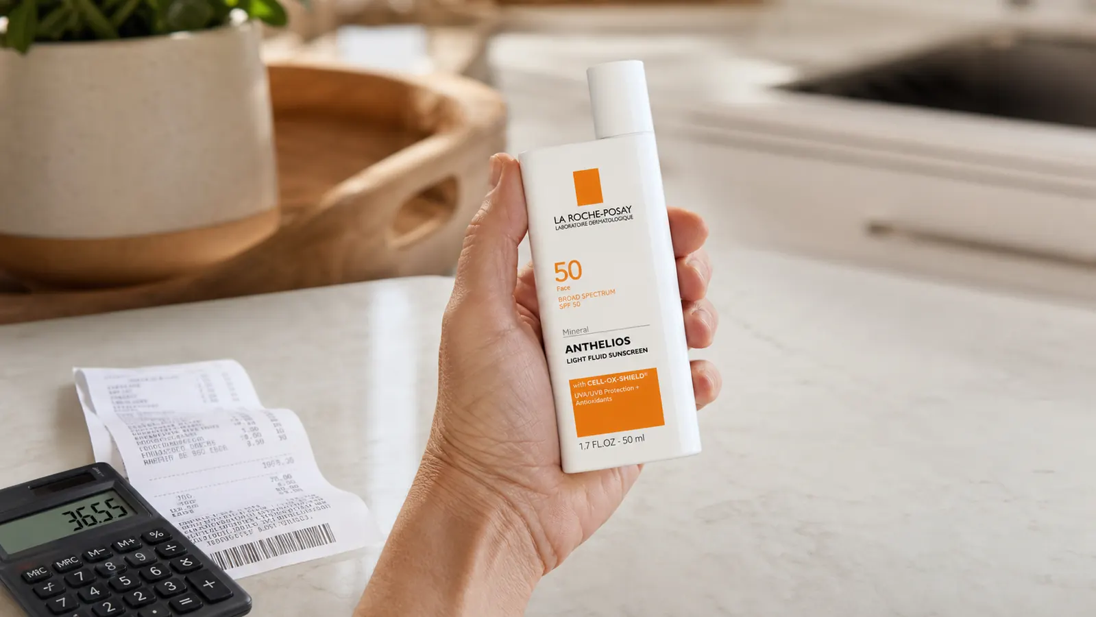 Close-up of a hand holding a slim tinted sunscreen fluid bottle next to a grocery store receipt and a calculator on a countertop