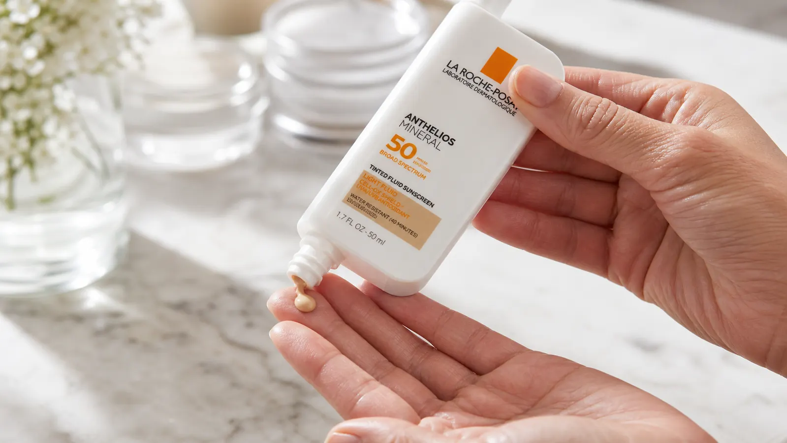 Close-up of a hand squeezing a small bead of tinted sunscreen fluid onto two fingertips above a bathroom sink