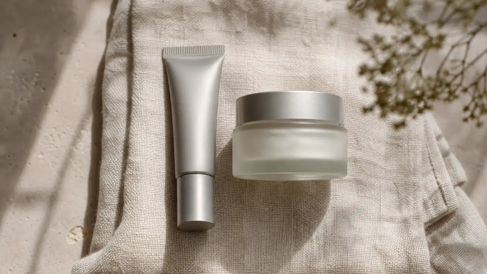 Two different eye cream containers, a metal tube and a round jar, side by side on a linen towel