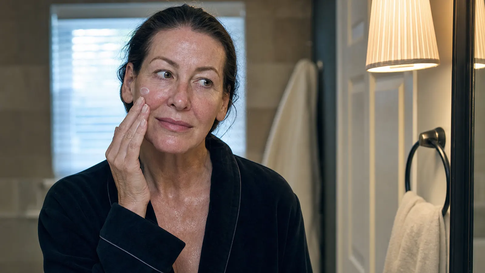 Woman patting moisturizer onto her cheek at a bathroom counter after cleansing, calm evening light