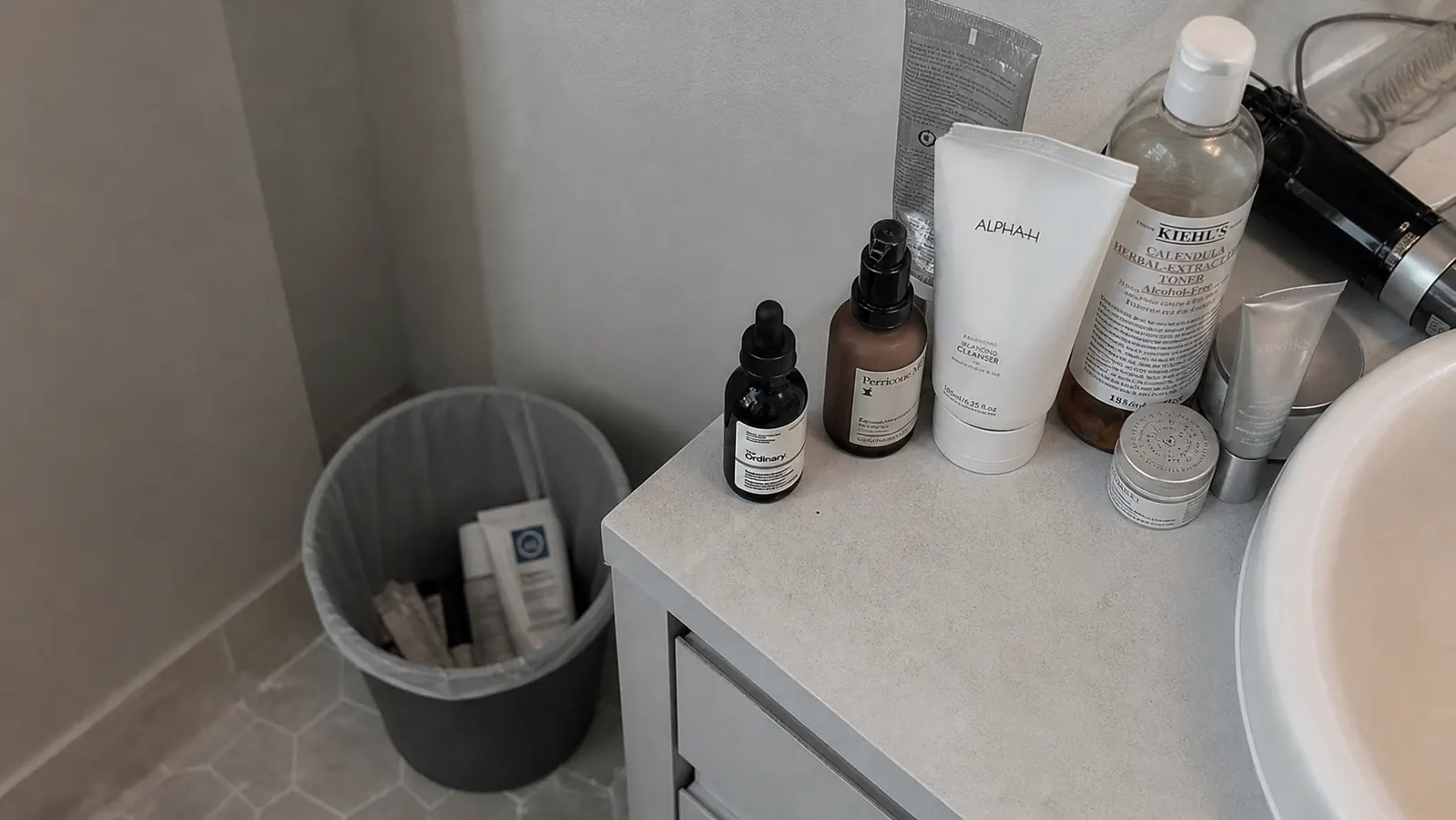 Cluttered bathroom counter with half-used cleanser bottles pushed to the back, next to a trash can with a couple of empty tubes in it