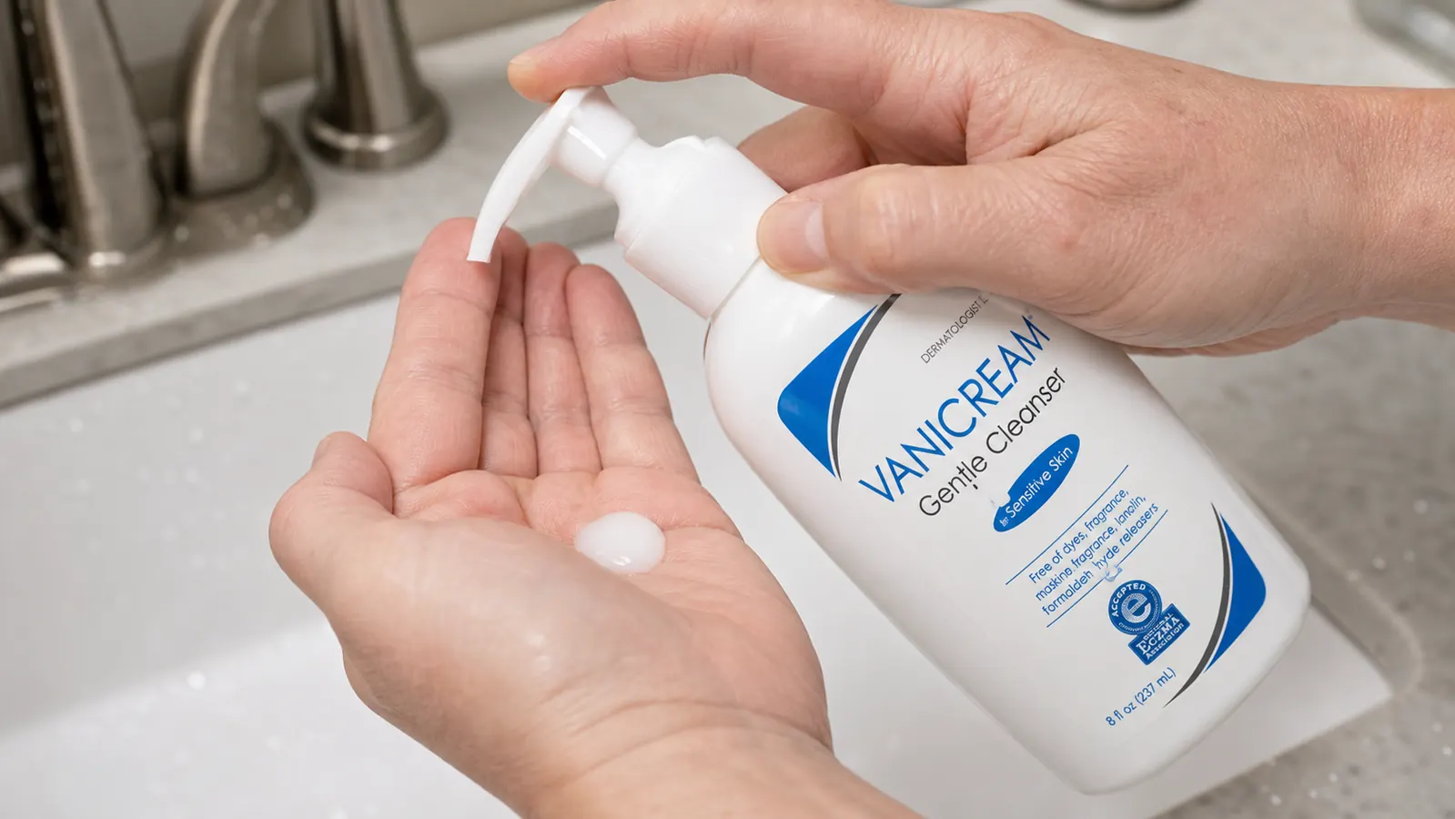 Close-up of a hand dispensing a small amount of white gentle cleanser into a palm above a bathroom sink