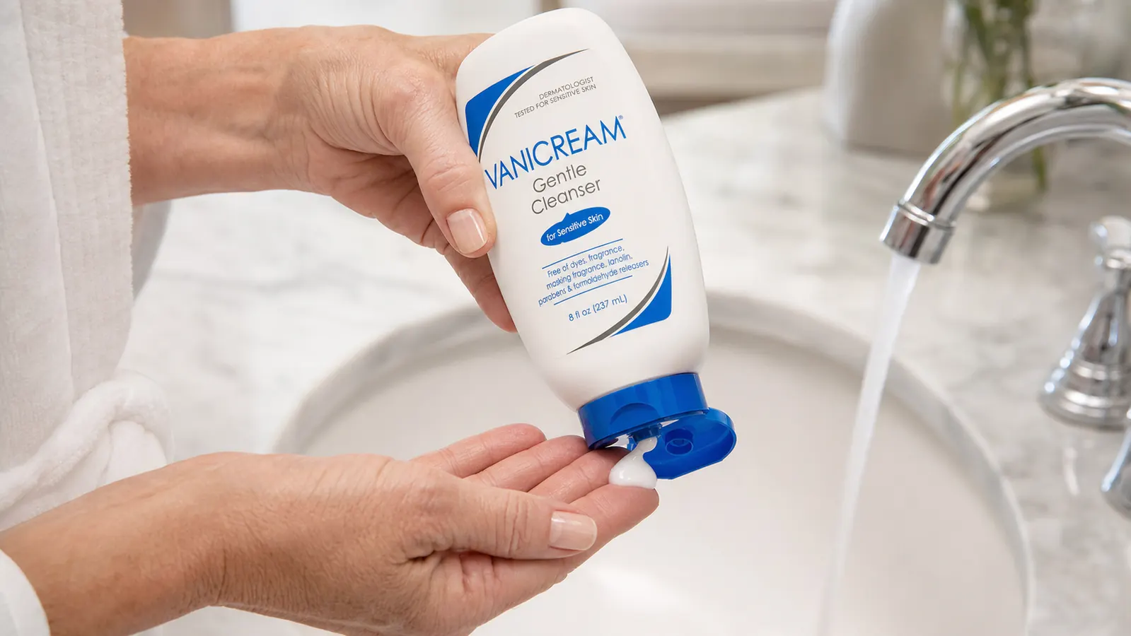 A hand squeezing a small amount of white lotion-textured cleanser onto fingertips over a bathroom sink